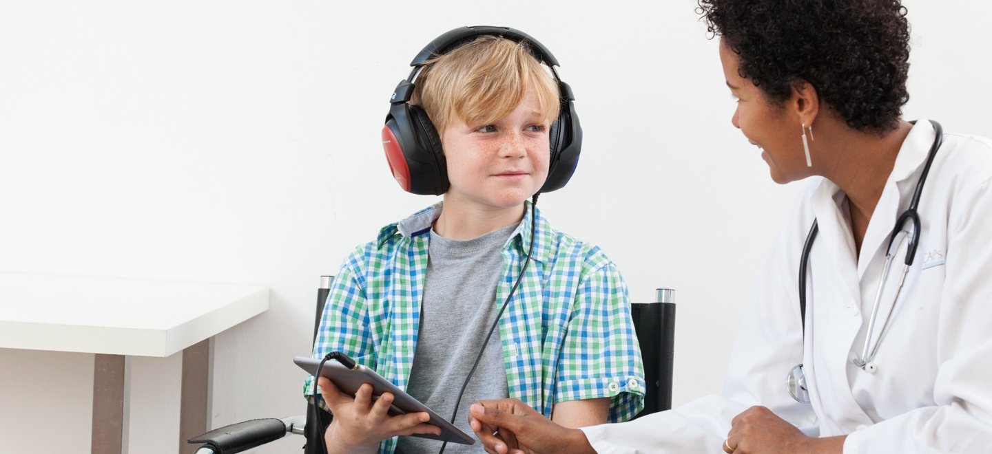 kid wearing audiometer headphones and taking the Jacoti hearing test in an iPad with help of an audiologist